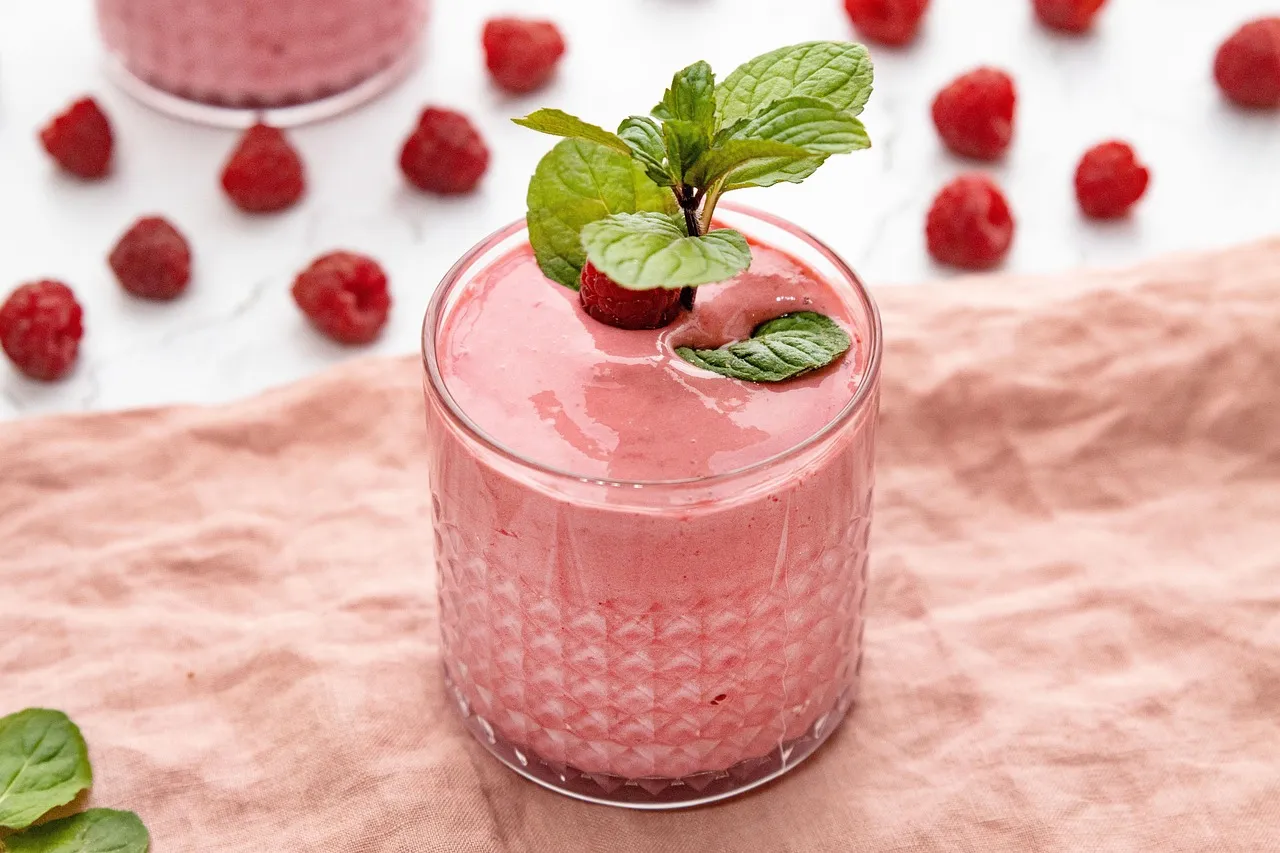 Vaaleanpunainen vadelmasmoothie lasissa, ympärillä tuoreita vadelmia ja mintunlehtiä